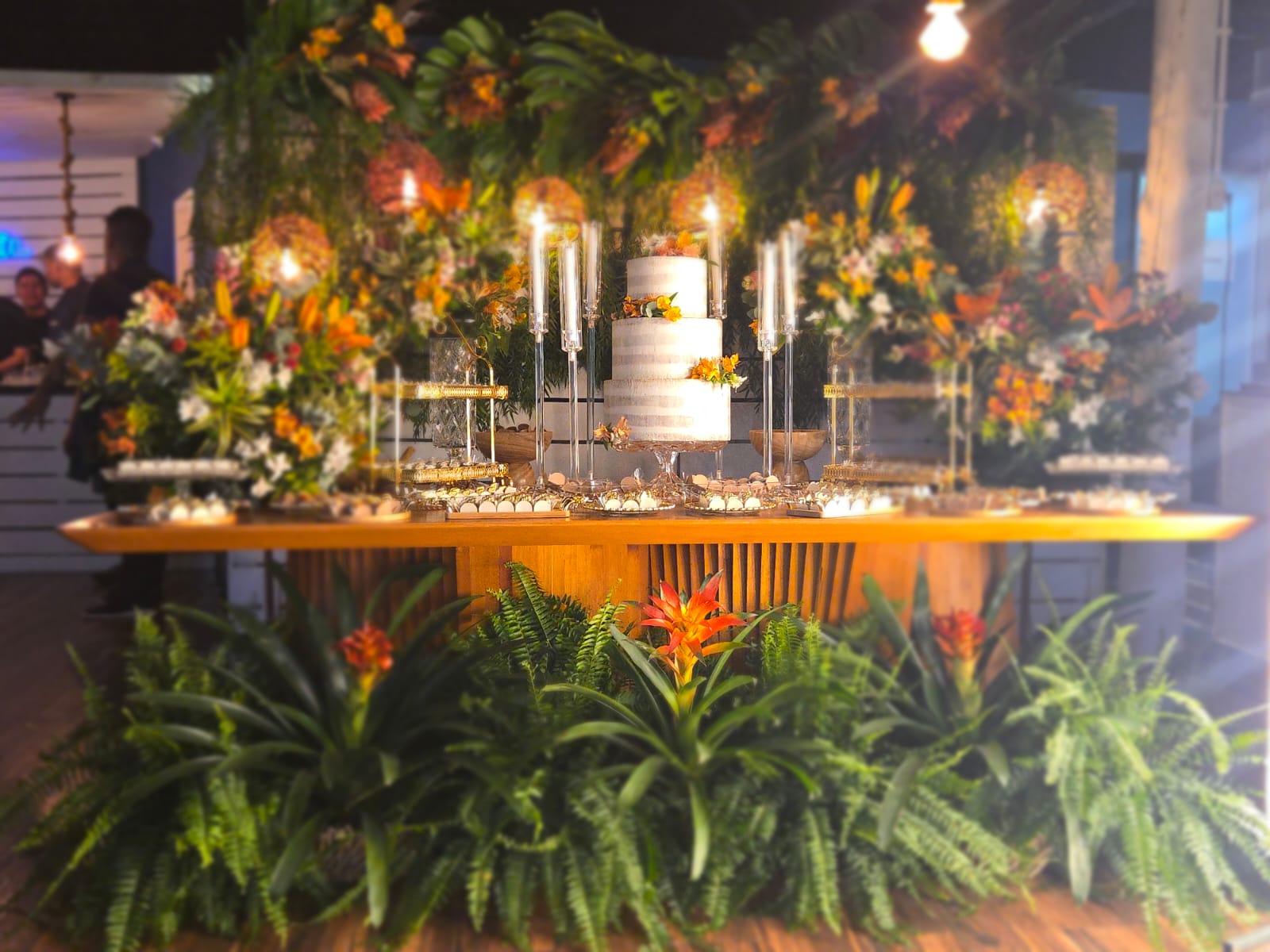 Mesa de doces com bolo branco e arranjos tropicais